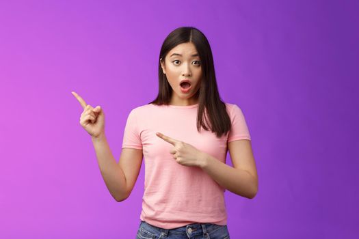 Shocked upset speechless cute asian girl react stunned friend got expensive car, drop jaw astonished, look camera full disbelief, pointing left, discuss astonishing news, stand purple background.