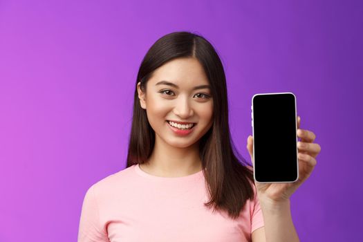 Close-up attractive pleasant female shop assistant present new phone app, hold smartphone show telephone screen, introduce application on display, stand purple background delighted.