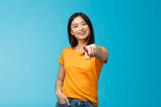 Lucky winner. Cheerful charismatic asian cute urban girl stand yellow t-shirt smiling friendly pointing finger camera choosing, picking person, inviting you team, stand happy blue background.
