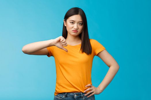 Moody unsatisfied cute asian girl picky, disliking bad restaurant food smirking displeased, grimacing disappointed show thumb down, shaking head negative judgement, stand blue background.