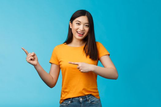 Upbeat friendly asian cute girl with dark haircut lively pointing left introduce awesome new copy space, smiling broadly toothy happy grin, stand blue background joyful, recommend link.