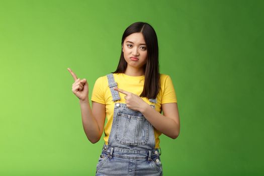 Disappointed uninterested asian gloomy girl pointing fingers left, grimacing sulking displeased, unwilling take part event, stare reluctant unimpressed, pose green background. Copy space