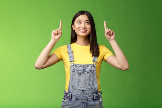 Dreamy hopeful cute asian girl look pointing up amused, smiling delighted, contemplate interesting object, enjoy stargazing, grinning satisfied, gazing top promo, stand green background.