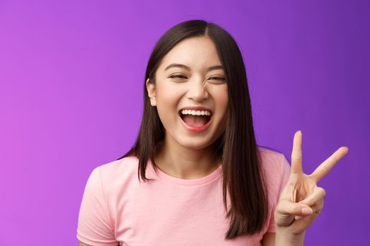 Headshot sincere carefree laughing asian girl having fun, show peace victory sign, joyfully look camera, express enthusiasm and happiness, enjoy funny friendly company, stand purple background.