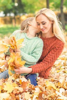 Happy young caucasian mother and little daughter holding autumn yellow leaves sitting and kissing at the park