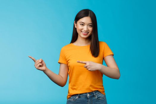 Friendly outgoing good-looking asian woman showing promo, pointing left indicating fingers advertisement, give advice, smiling confident, pleasantly help pick clothes, stand blue background.