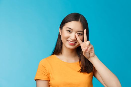 Positive cute and silly asian girlfriend posing photograph lovely smiling, show peace victory sign near face grinning friendly, carefree enthusiastic mood, stand blue background in yellow t-shirt.