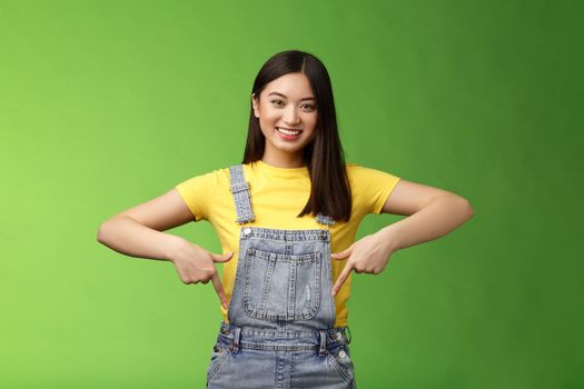 friendly outgoing cute asian young woman show fingers down, introduce new promo app, smiling joyfully sharing link, recommend product using brand, grinning toothy, green background. Copy space