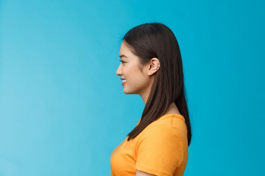 Profile shot asian cute woman perfect skin, turn left, smiling joyfully, pose near blue background, standing queue, awaiting coffee take-away, relaxed chatting girlfriend, wear yellow summer t-shirt.