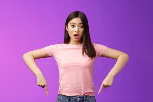 Shocked upset cute asian surprised girl, drop jaw shook disappointed, gasping disappointed pointing down amazed, indicating bottom advertisement, judging, negative opinion, purple background.
