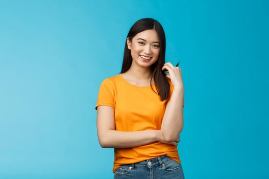 Cheerful asian girl with dark short haircut touching hair strand smiling lively, cross one arm chest, laughing, carefree talking casually friends, discuss university lifestyle, blue background.