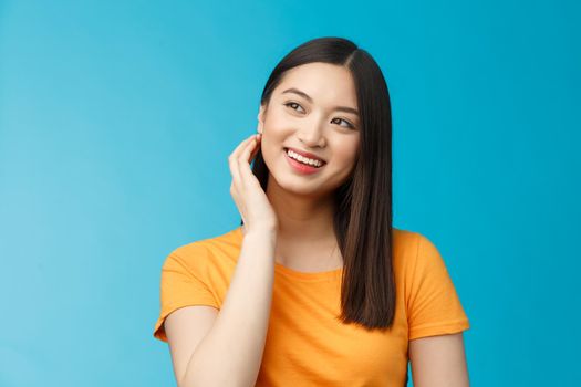Tender lovely coquettish modern asian woman touch hair lovely look aside, smiling joyfully, feel satisfied after good haircut, deal with nasty pimples, get rid acne delighted, stand blue background.