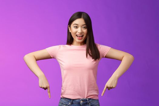 Lively enthusiastic asian brunette female excited telling about awesome promo, pointing down surprised, grinning joyfully recommend good advertisement, indicating bottom advertisement.