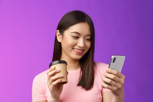 Close-up tender friendly asian female blogger checking social media drinking take-away coffee, smiling joyfully look telephone screen, reading interesting article, texting, purple background.