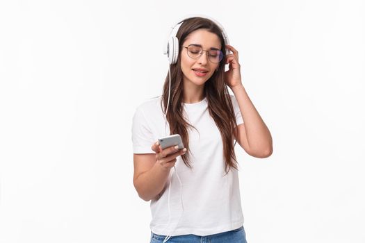Technology, lifestyle and music concept. Portrait of pleased and relaxed young woman vibing and chilling as listening favorite song in headphones, hold mobile phone, smile and close eyes.