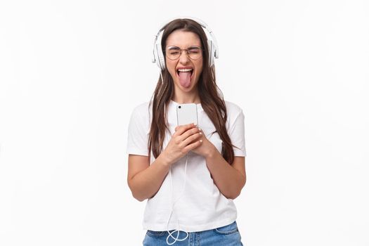 Technology, lifestyle and music concept. Portrait of carefree happy and enthusiastic woman enjoying awesome song, listening in headphones hold smartphone show tongue, white background.