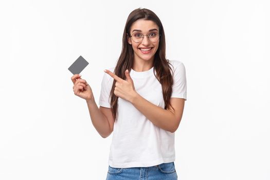 Portrait of excited, happy young woman talking about her bank, pointing at credit card and smiling camera, advice use this for non-cash payment, purchasing goods online, white background.