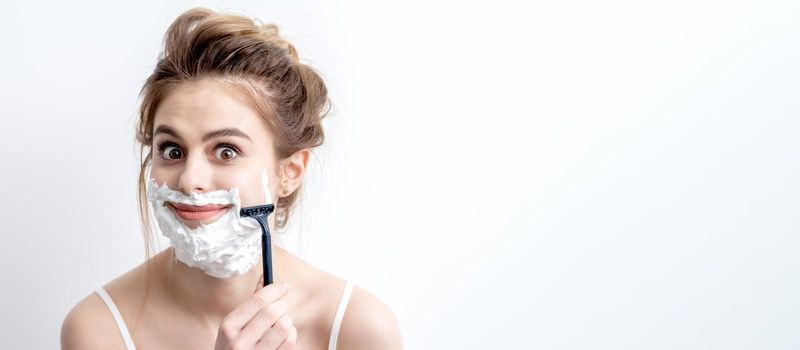 Beautiful young caucasian woman shaving her face by razor on white background. Pretty woman with shaving foam on her face