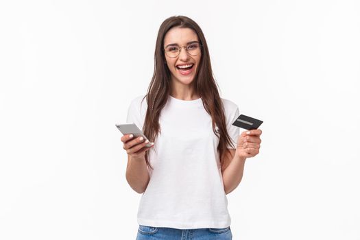 Portrait of happy smiling pretty woman in glasses holding credit card, mobile phone, laughing at camera easily paying for her purchase online store, buy things with application, white background.
