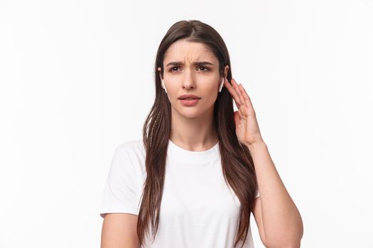 Close-up portrait of serious displeased young woman having troubles with using wireless earphone, have bad quality of sound, touching earbud to turn on song, frowning perplexed, white background.