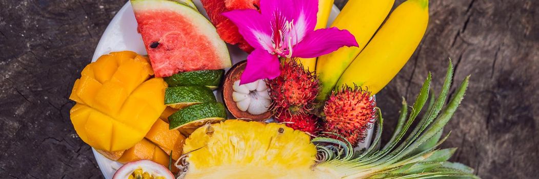 Colorful tropical fruits on big plate. On rustic wooden background. Top view. BANNER, LONG FORMAT