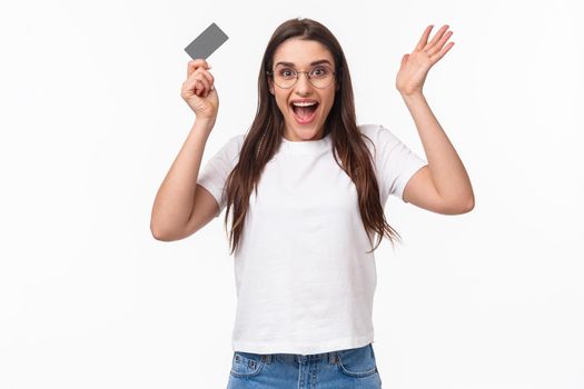 Waist-up of enthusiastic, happy surprised young woman reacting to wonderful news, raising hands up in amazement, holding credit card, receive salary, got her first paycheck, hurry to order something.