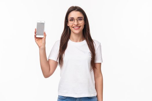 Communication, technology and lifestyle concept. Portrait of happy smiling friendly-looking girl in glasses, t-shirt, showing smartphone display, mobile phone application, recommend upload it.