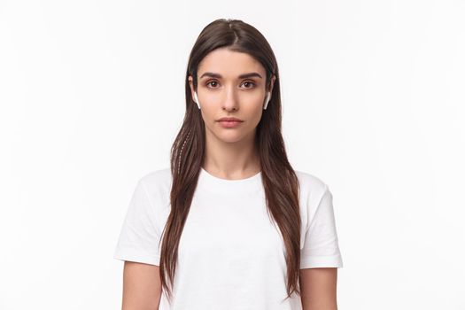 Close-up portrait of serious-looking attractive caucasian woman in wireless earphones, looking at camera with relaxed no emotions face, standing white background. Copy space