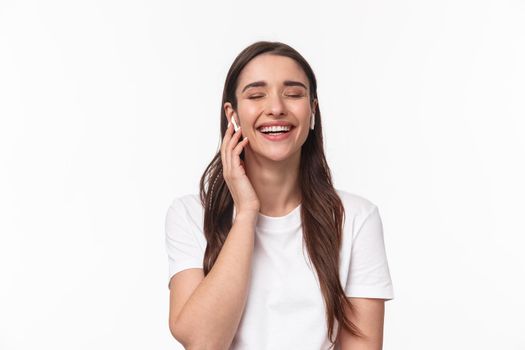 Close-up portrait of adorable caucasian woman, smiling and laughing with closed eyes, touching wireless earphones, having phone conversation with headphones, white background.