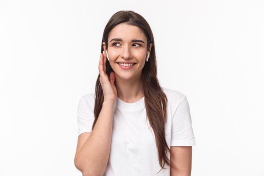 Close-up portrait of attractive young caucasian female in t-shirt, listening favorite song, using wireless earphones, smiling and look away dreamy, touching headphone to change song in playlist.