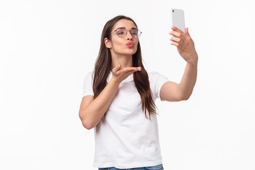 Communication, technology and lifestyle concept. Lovely attractive woman in glasses, sending air kiss to her followers online live stream with app, taking photo with filters, hold mobile phone.