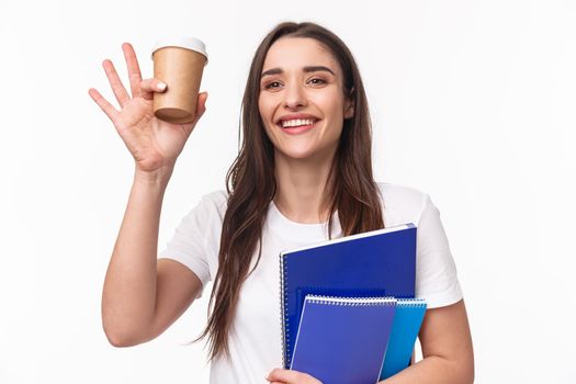 Education, university and studying concept. Close-up portrait of cheerful, friendly beautiful woman drinking coffee, saying hi to classmate in campus, holding school supplies, books and notebooks.