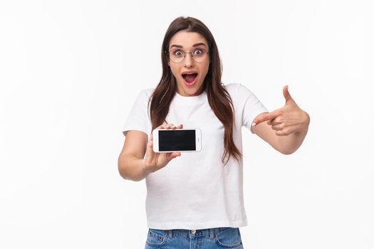 Communication, technology and lifestyle concept. Portrait of surprised and excited young woman in glasses hurry up with announcement of awesome new application, pointing at mobile phone.