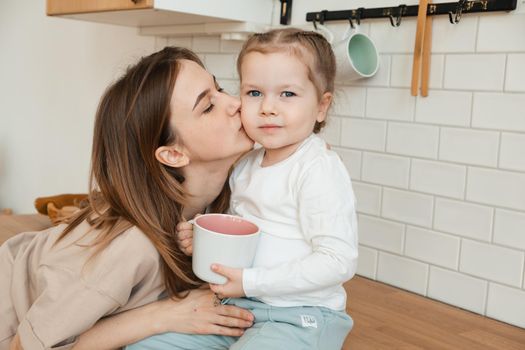 Mother kissing her daughter at home in kitchen, breakfast. Little girl looking at camera