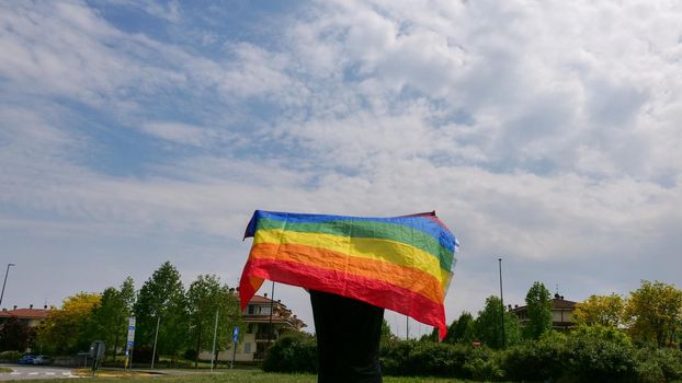 Bisexual, girl, lesbian, woman, transgender, homosexsual holding in hand a rainbow LGBT gender identity flag on sky background with clouds on a sunny day and celebrating a gay parade in pride month