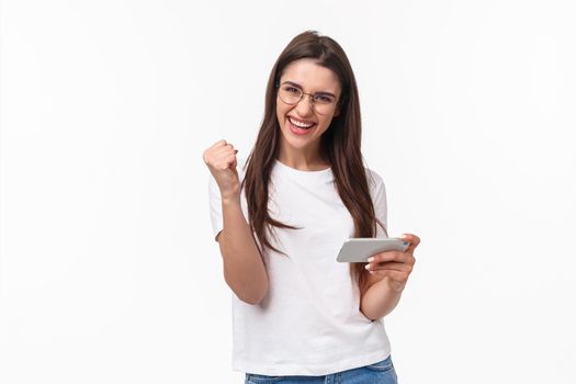 Communication, technology and lifestyle concept. Portrait of successful cheerful lucky girl winning prize in game, finished hard level, fist pump after win on mobile phone app, white background.