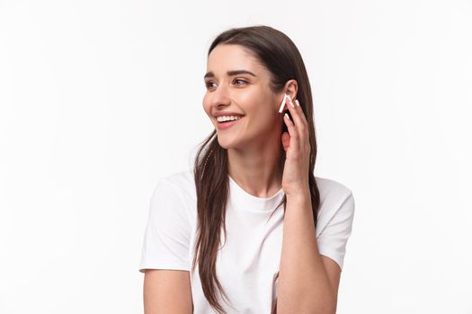 Close-up portrait of happy charming young woman with long hair, looking away smiling and laughing joyfully, changing song touching her wireless earbud, stand white background.