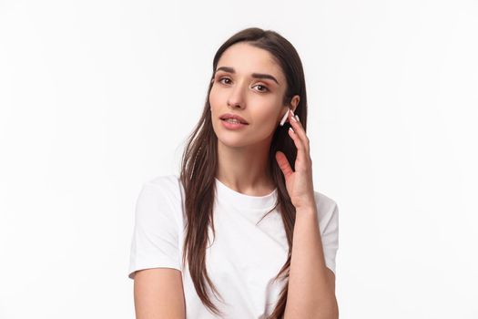 Close-up portrait of feminine attractive young woman in t-shirt, touching wireless earbud, listening music in headphone, looking camera, turn-on podcast or relaxing playlist, white background.