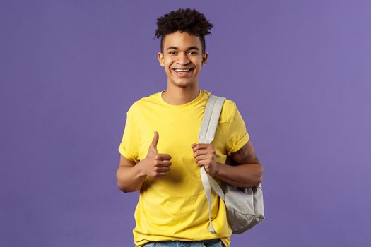Back to school, university concept. Portrait of cheerful handsome male student describe his summer vacation to classmates, show recommendation, thumbs-up approval, hold backpack.