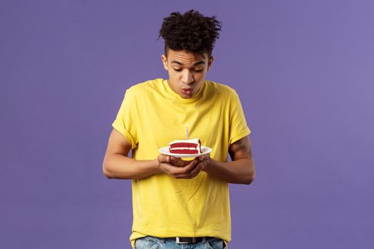 Portrait of cute excited young guy with dreads and tattoos, making wish on birthday, celebrating party, blowing candle on b-day cake, standing purple background dreaming. Holidays concept