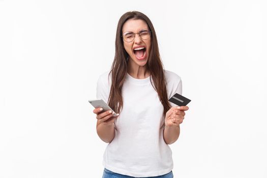 Portrait of uneasy annoyed and angry young brunette female in t-shirt yelling pissed-off as checking bank account, have no money on credit card, or it expired, hold mobile phone, white background.