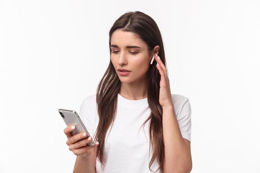 Close-up portrait of modern young woman in t-shirt, using wireless earphones listening music or podcasts, picking song in mobile phone, texting friend or calling person, white background.