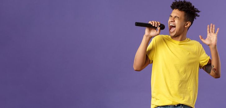 Portrait of passionate carefree young hispanic singer with dreads and tattoos, reaching highest note in song, raising hand up singing loud at microphone with closed eyes, purple background.