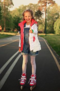 little girl on roller skates makes a gesture thumb up in the Park on a Sunny day