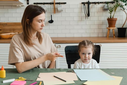 Cute angry little preschool girl doing creative homework with her mom while sitting at the table in the living room and arguing. Problems in making crafts for kindergarten