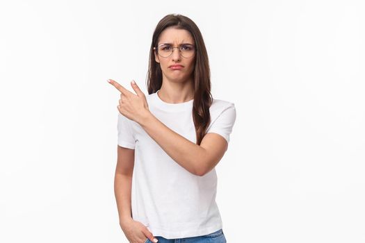 Waist-up portrait of moody, disappointed and complaining cute brunette woman in glasses, frowning and grimacing, sulking defensive, pointing finger upper left corner at something offensive.