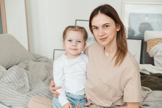 Portrait of happy loving single mother hugging cute little preschool daughter and sitting on bed looking at camera