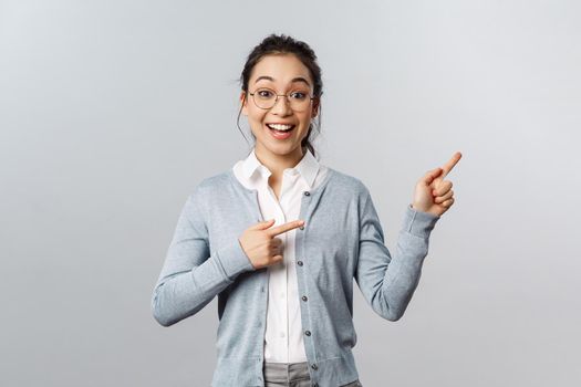 Upbeat excited mixed-race girl in glasses, asian woman teacher pointing fingers right, smiling broadly, found way to stay in touch with students during lockdown quarantine, standing grey background.