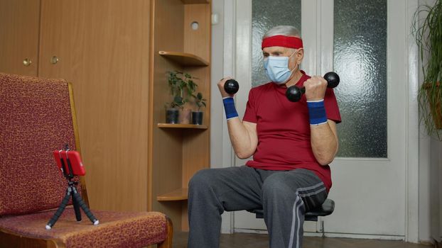 Retired senior man in medical mask doing workout, training, fitness, sport activity exercises at home. Elderly old grandfather making weight lifting dumbbells exercising during coronavirus lockdown