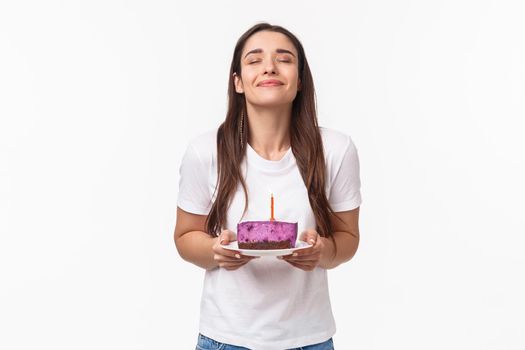 Entertainment, fun and holidays concept. Portrait of romantic beautiful young b-day girl celebrating throwing party, holding piece of birthday cake with candle, making wish with smile and closed eyes.